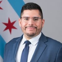 Headshot of a man in a suit and tie and wearing glasses, in front of the Chicago flag.